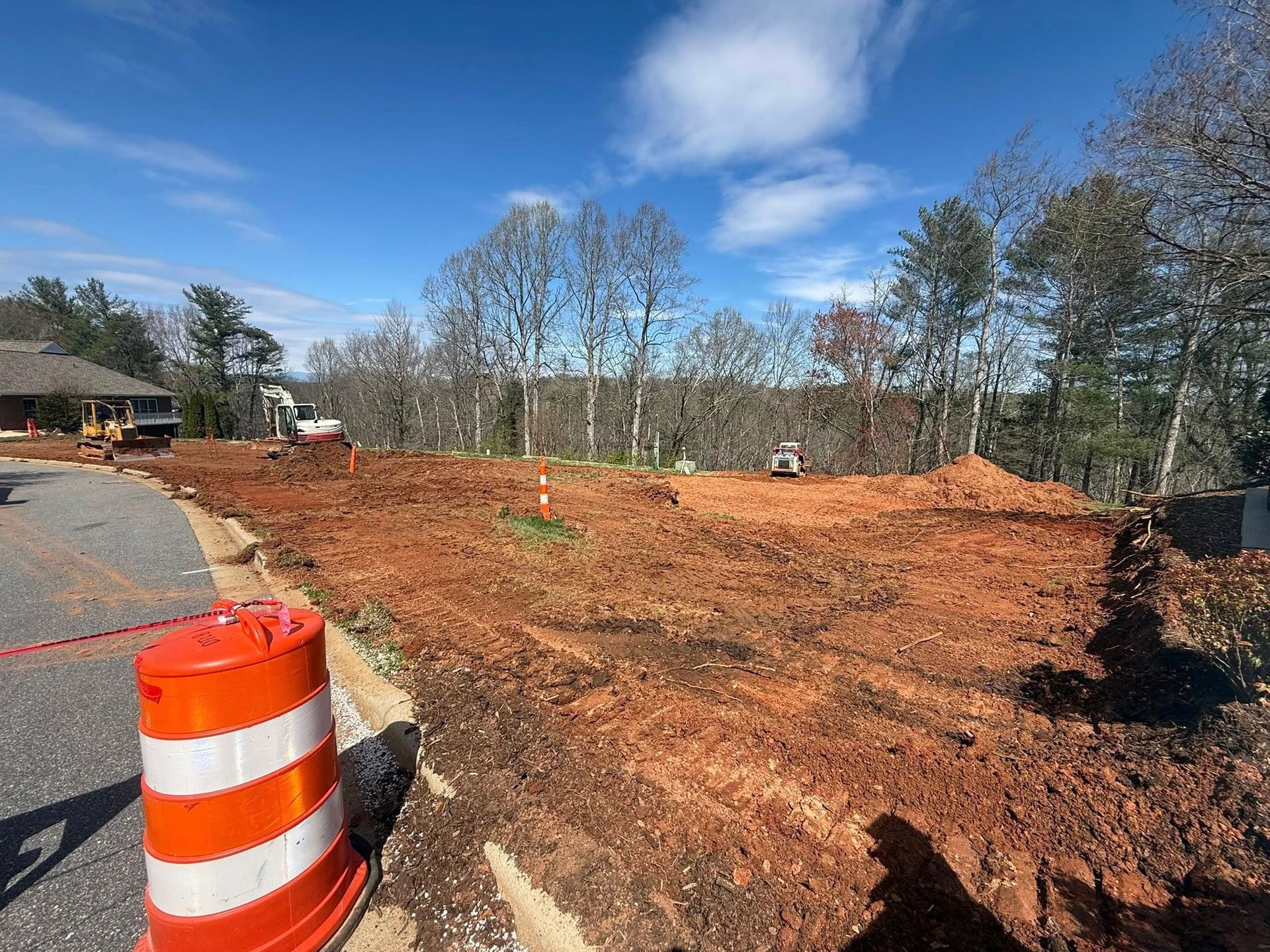 Construction site with orange traffic barrel, dirt grading, and trees under a blue sky
