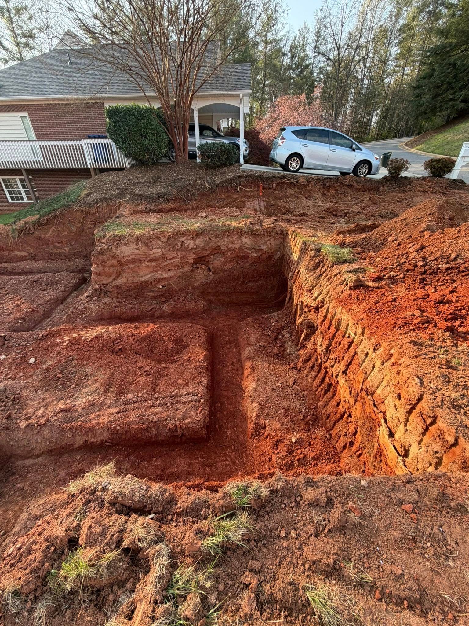 Large excavated dirt hole in a front yard, with a house and white car in the background.