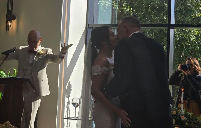A bride and groom kissing during their wedding ceremony while a man stands at a podium.