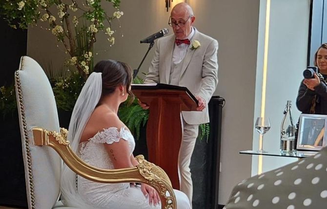 A man is giving a speech at a wedding ceremony while a bride sits in a chair.