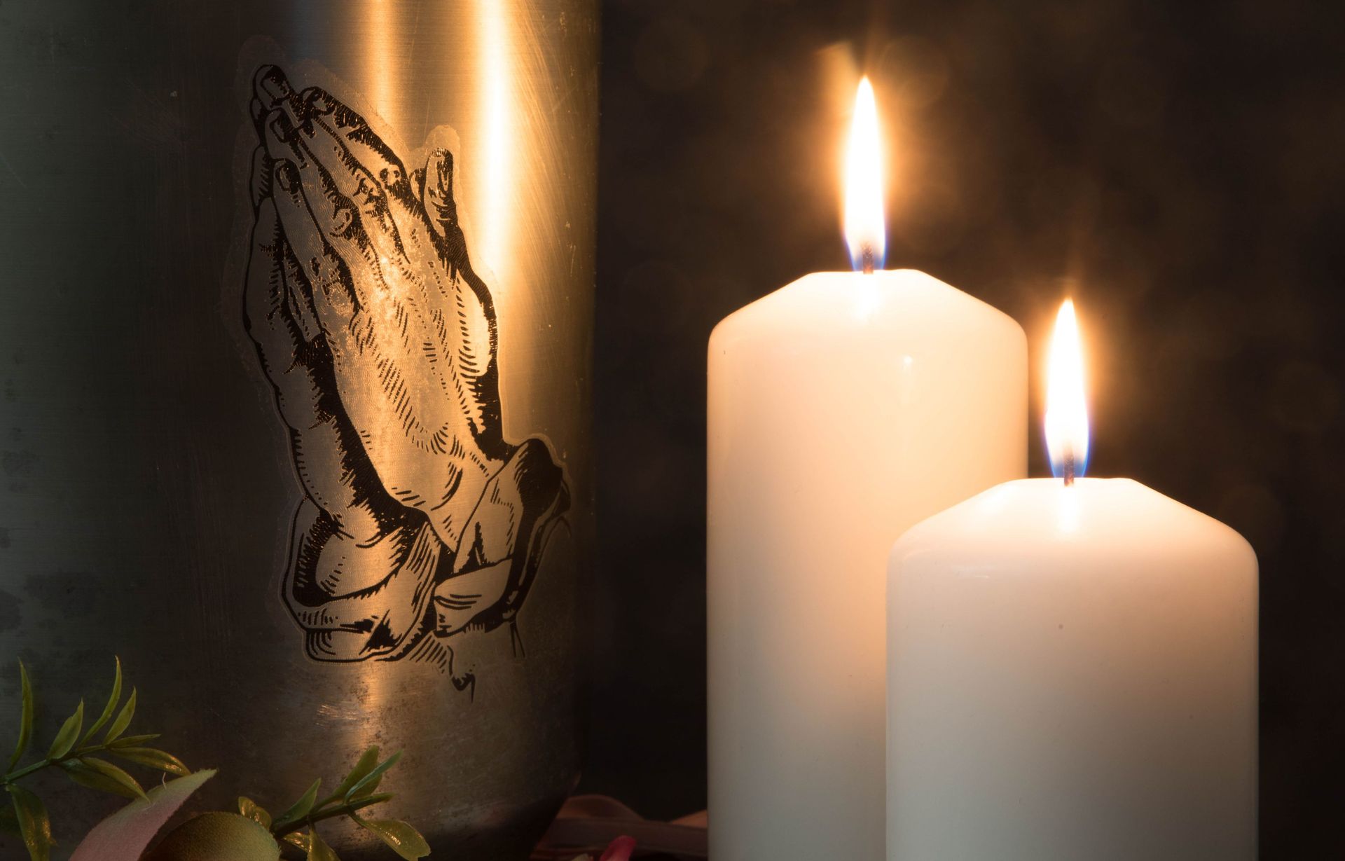 Two candles are lit in front of a metal urn with praying hands on it.