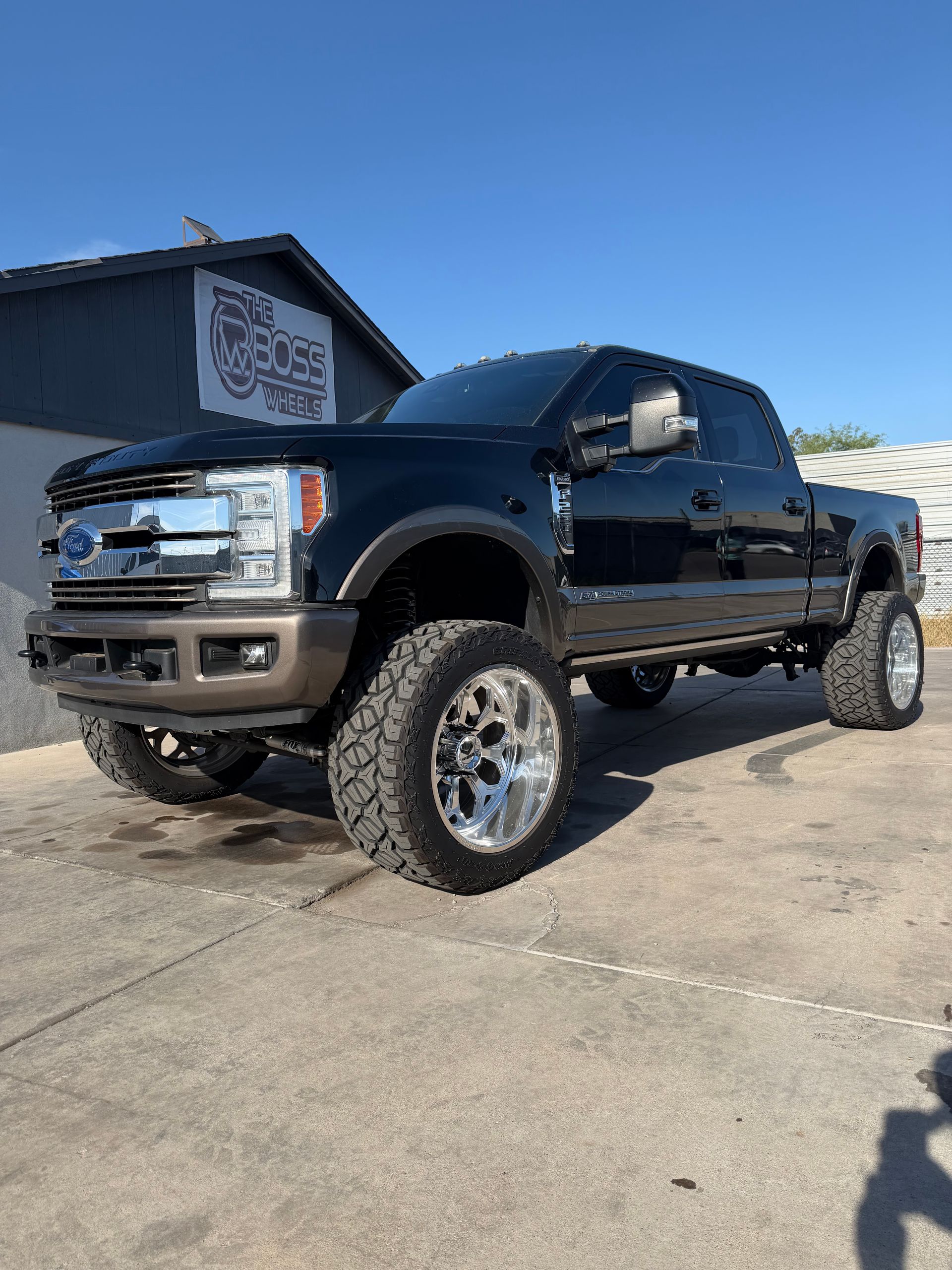 Black lifted pickup truck parked on a concrete lot beside a building under a clear blue sky.