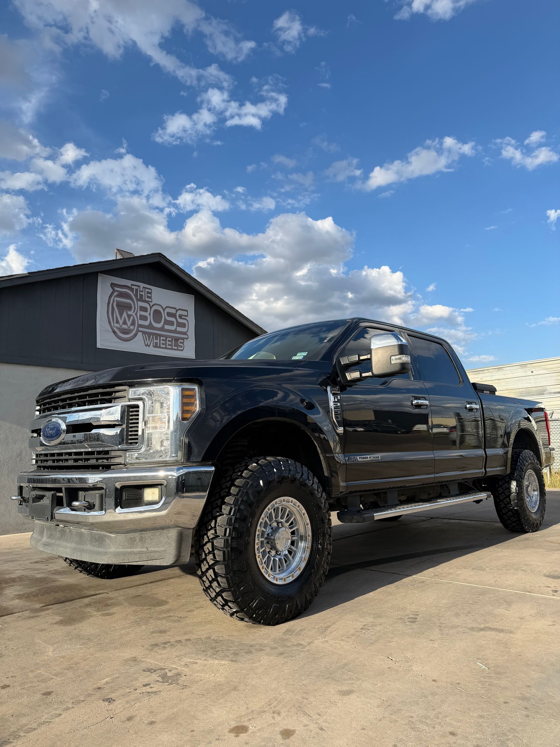 Black lifted pickup truck parked on a dirt lot under a blue sky with clouds