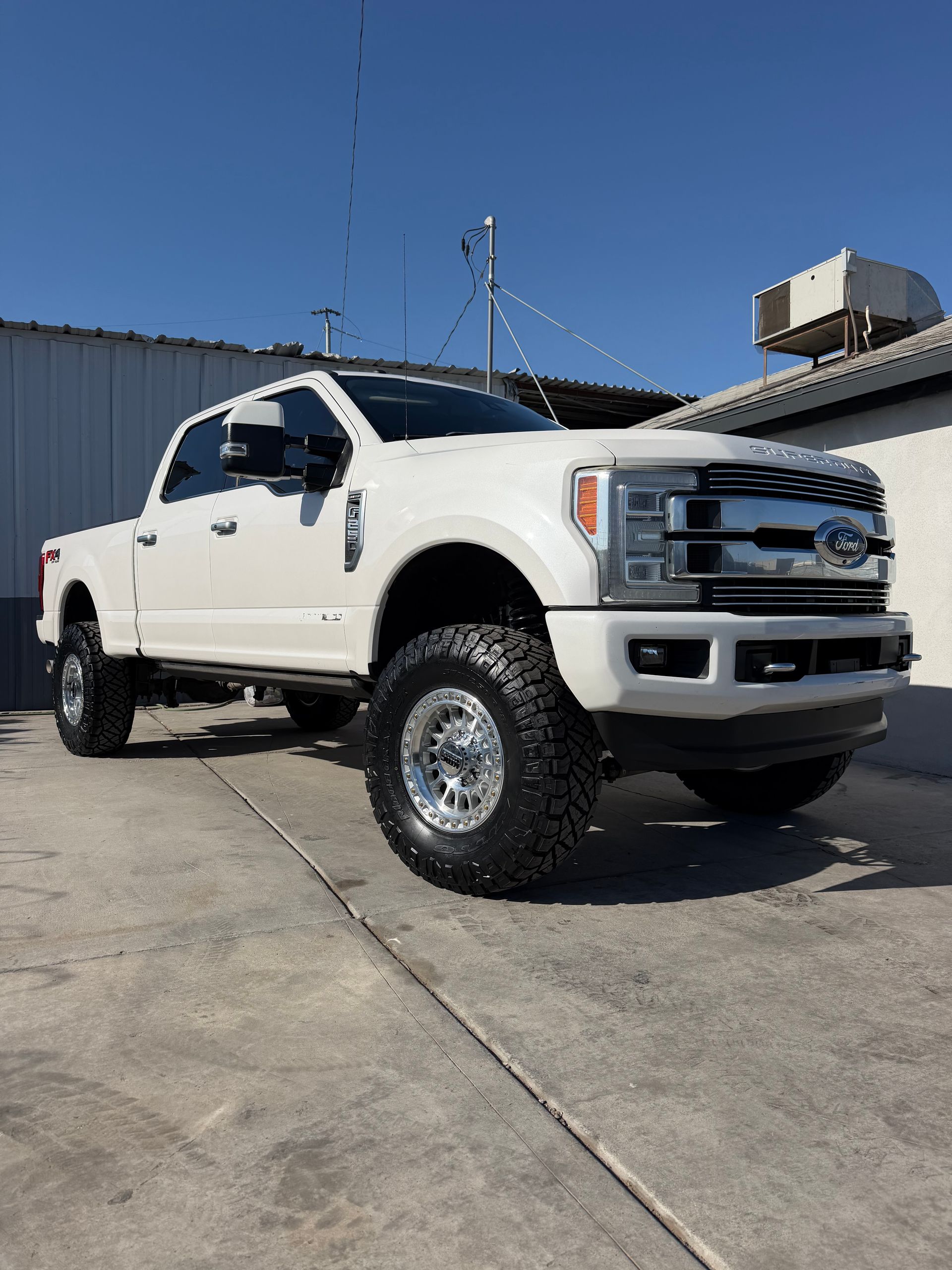 White lifted Ford pickup truck parked on pavement beside an industrial building under a clear blue sky