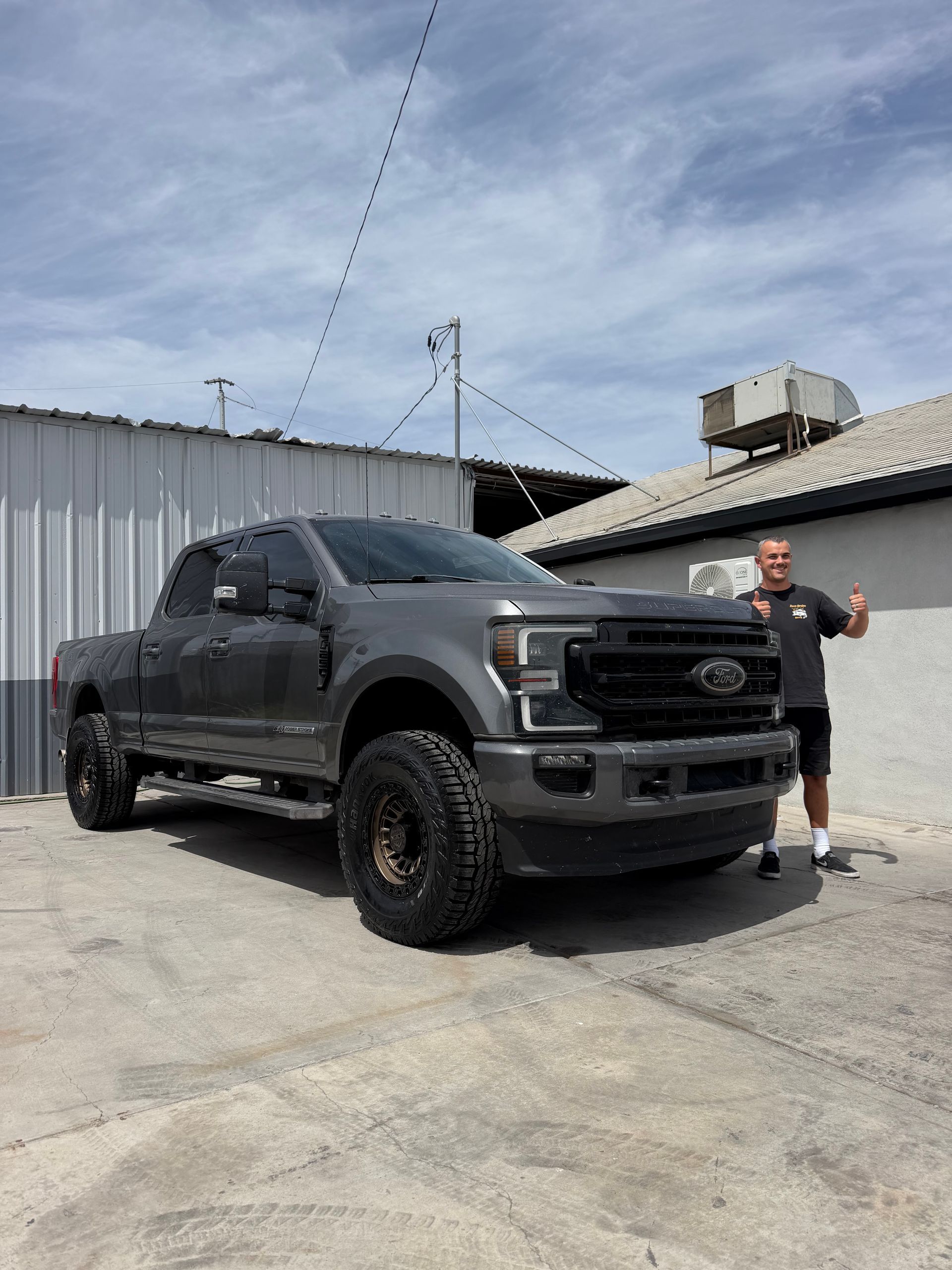 Lifted dark gray pickup truck parked outdoors beside a person in a black shirt and shorts, waving.
