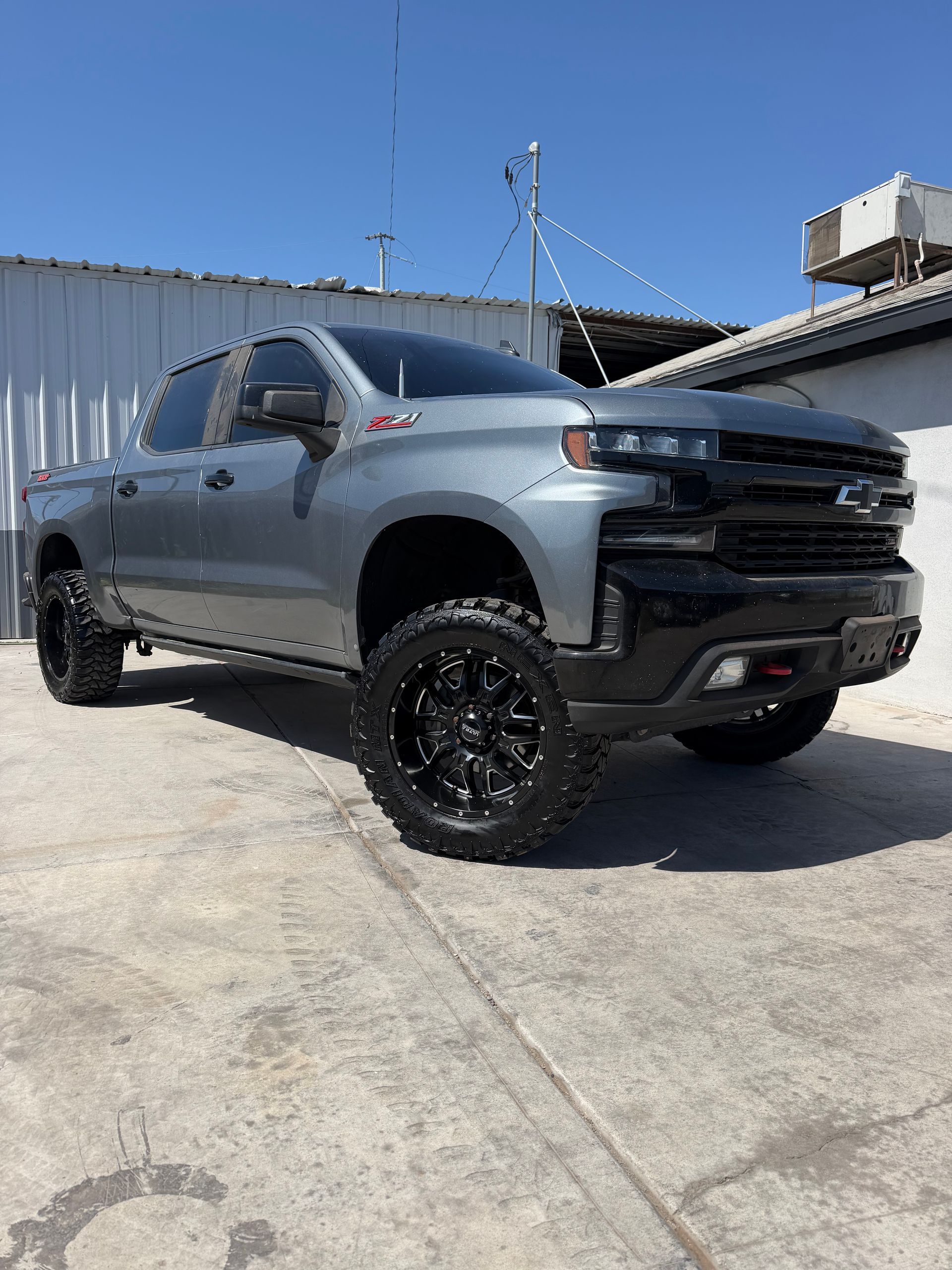 Gray lifted pickup truck with black wheels parked on a concrete lot beside a building