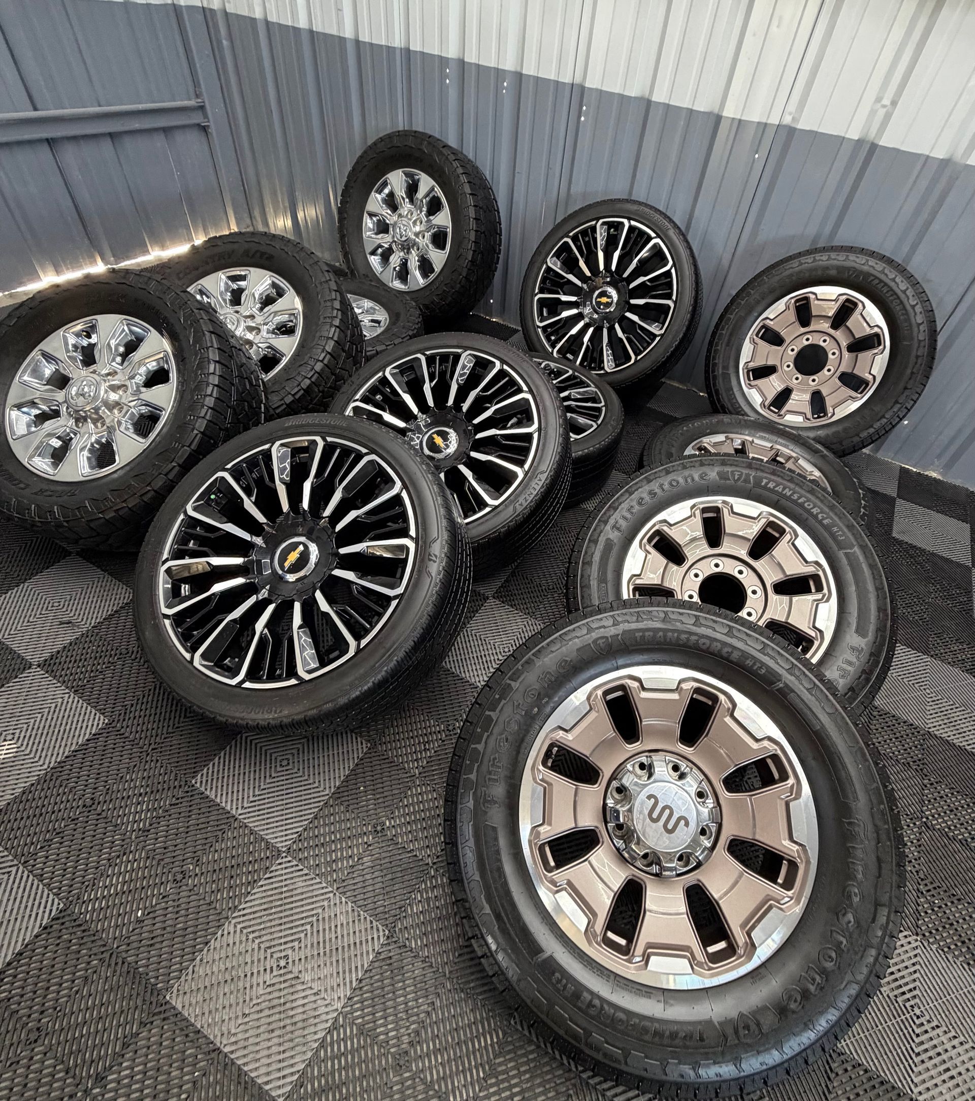 Assorted car tires mounted on alloy rims, stacked on a checkered floor against a corrugated wall.