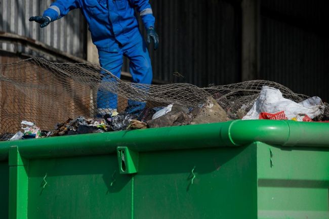 Person in blue work suit on a green dumpster, securing netting over trash.