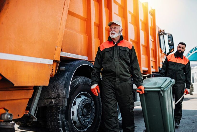 two men wheeling a trash bin next to a large orange dump trush