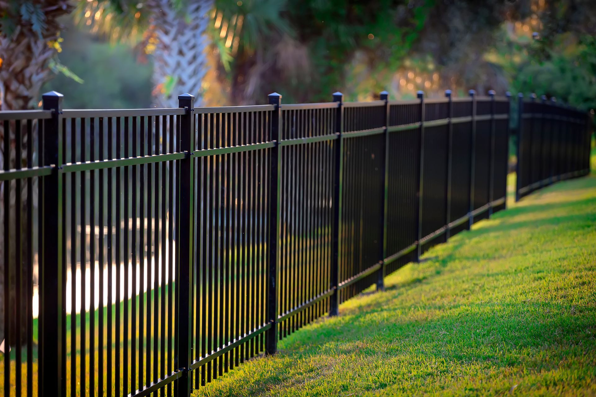 A black metal fence stretches across a green lawn near palm trees and a pond in the background.