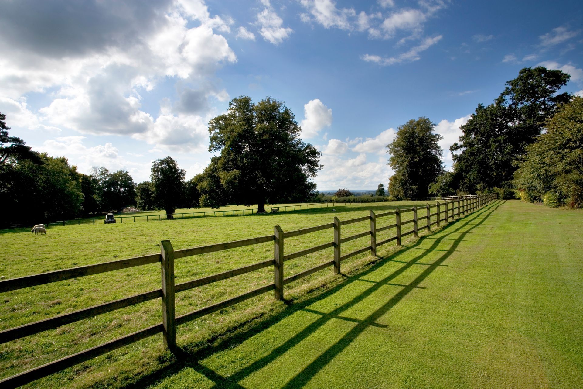 A rural landscape with a long wooden fence separating a lush green pasture from a mown lawn under a bright, cloudy sky.
