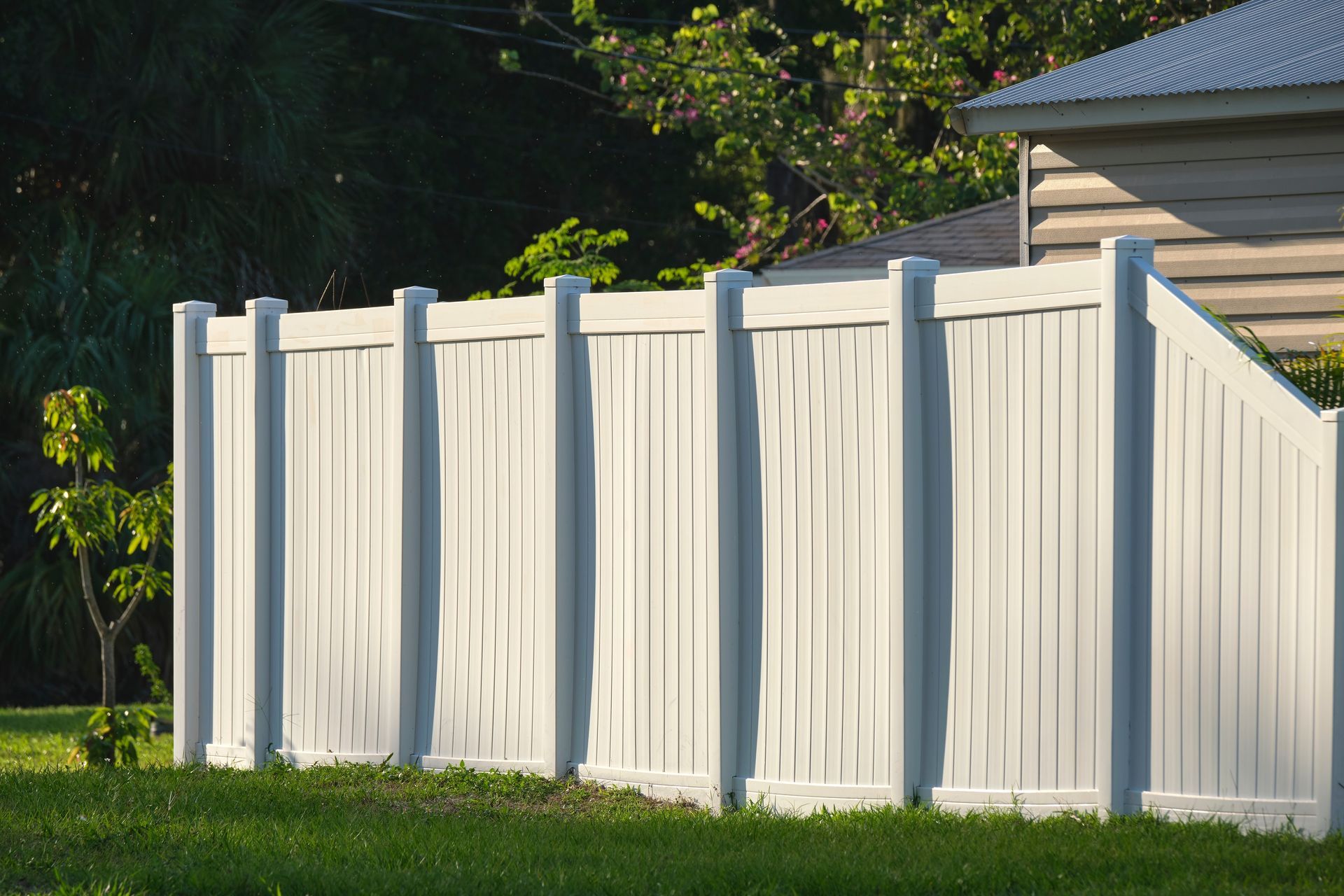 A white vinyl privacy fence stands in a grassy yard, with a partial view of a house and trees in the background.
