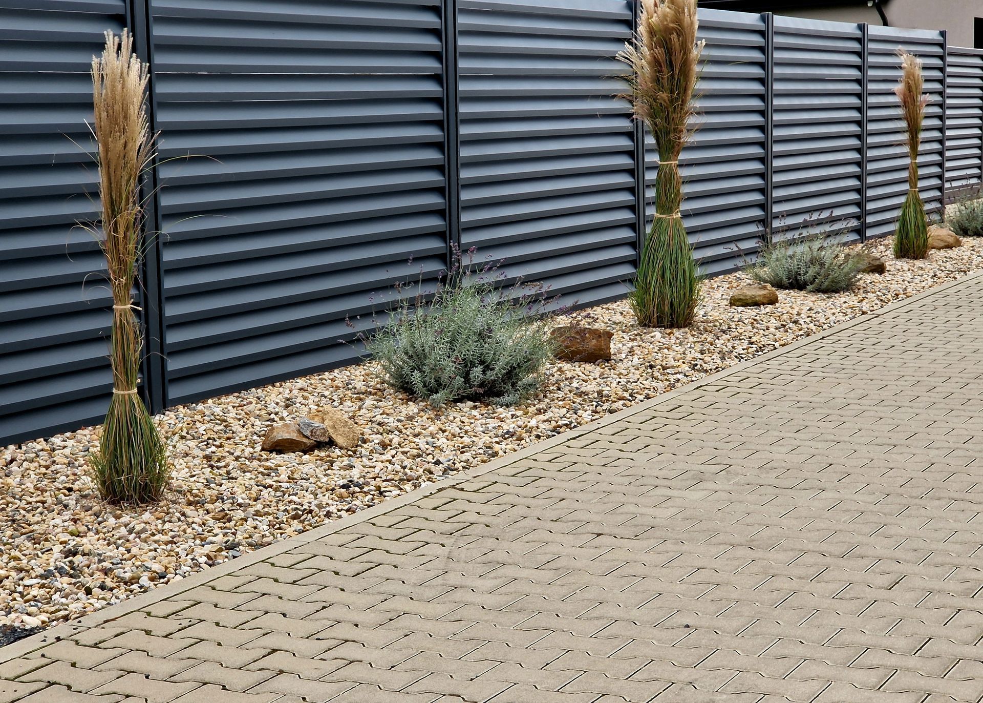 A modern, dark grey corrugated metal fence borders a paved walkway with decorative gravel, ornamental grasses, and stones.