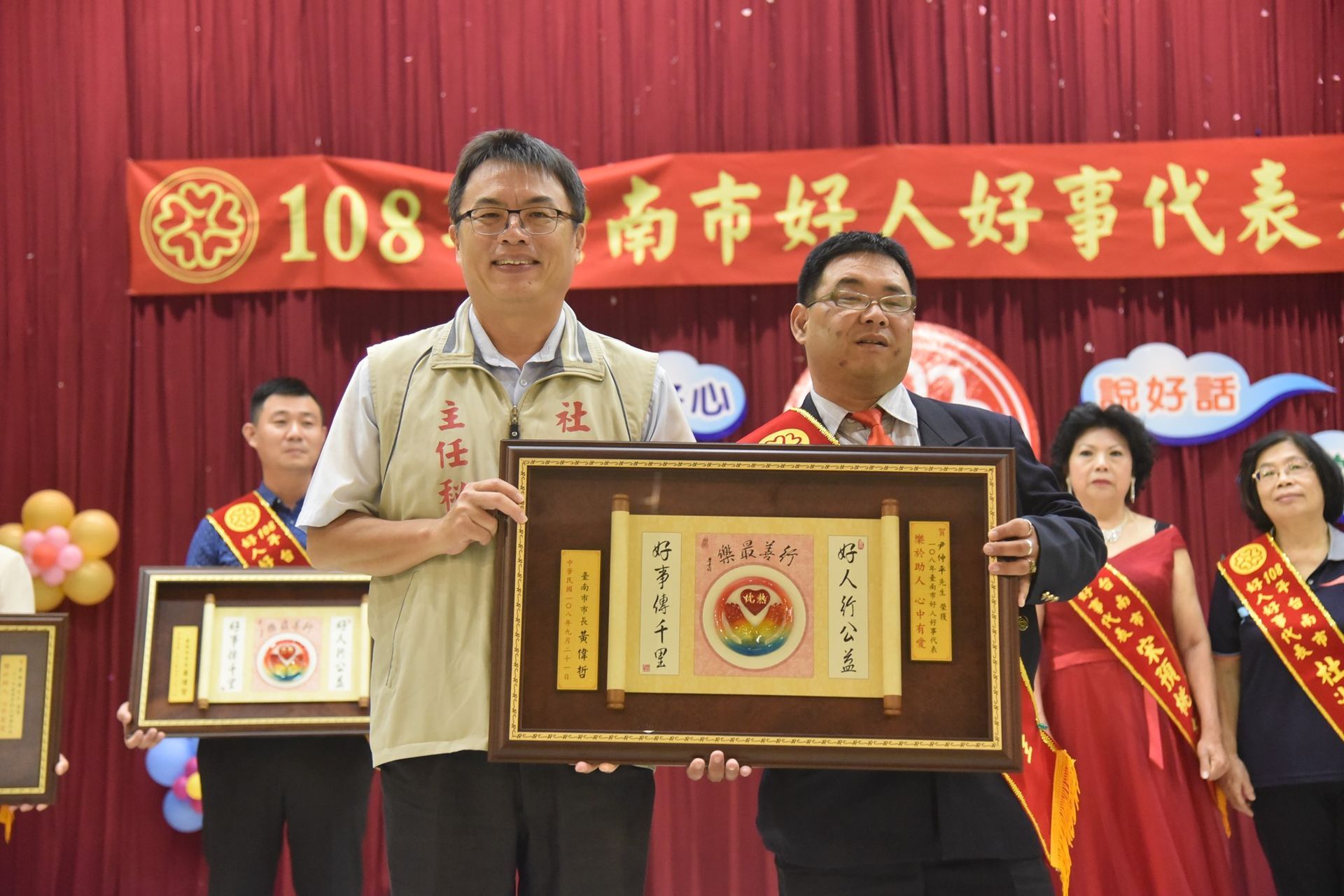 兩名男子手捧裱框獎項站在舞台上,在紅色背景前慶祝一場活動。