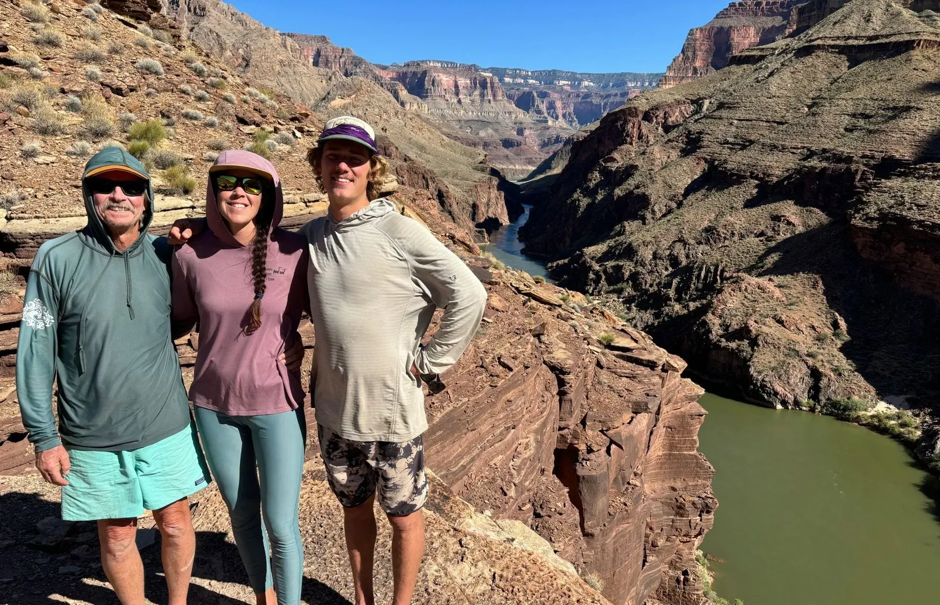 Three people pose on a canyon rim overlooking a river. They wear outdoor clothes, with hats and sunglasses.