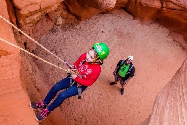 A young girl is hanging from a rope in a canyon while the father looks on