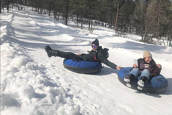 Two people snow tubing down a snowy hill, one lying back on a blue tube and one sitting upright.