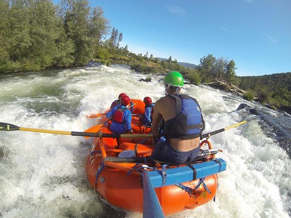People whitewater rafting in an orange raft on a fast, rocky river with trees and blue sky