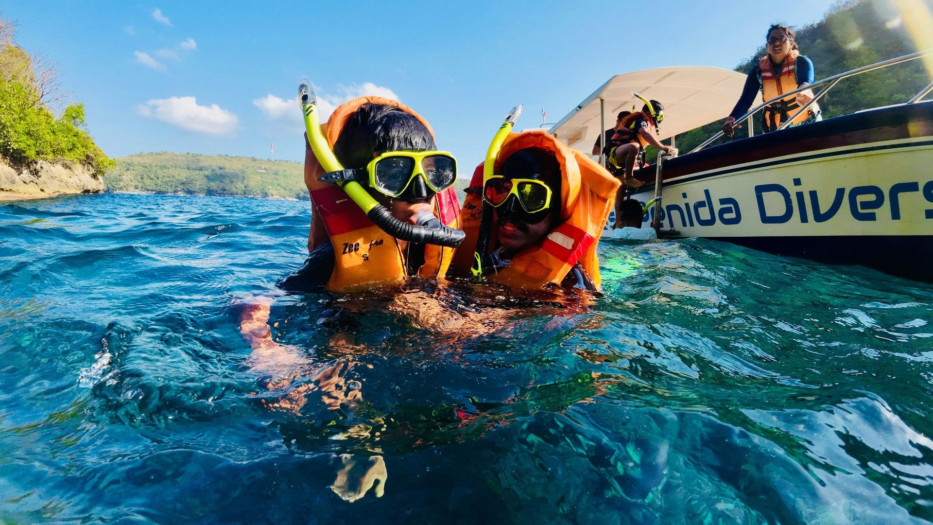 Two snorkelers in orange vests float in blue water beside a boat, near a tropical coastline.