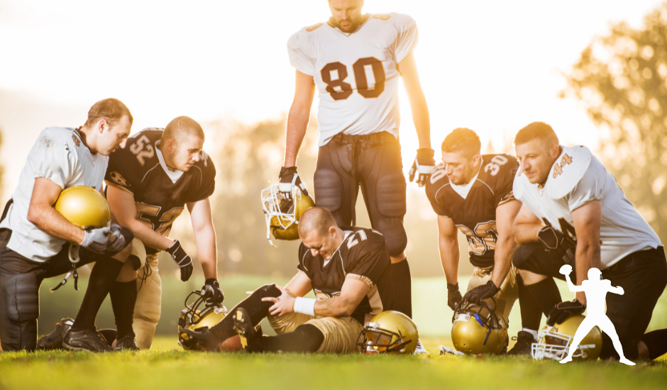 American football players huddle around an injured teammate on a sunny field. Several players in black and white uniforms help the player who is clutching his leg.
