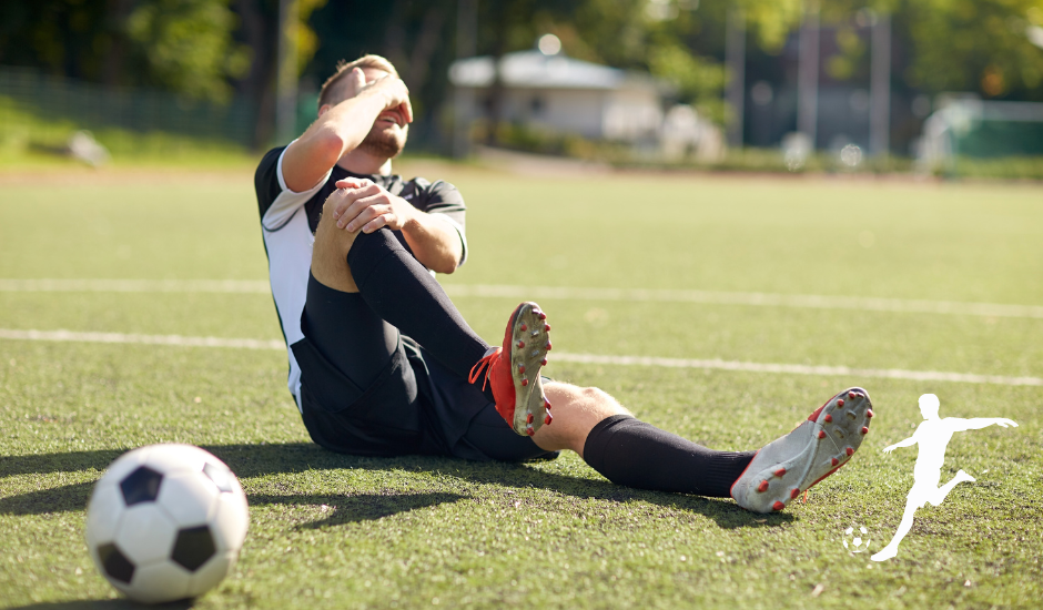 Soccer player sitting on turf, holding ankle in pain beside a soccer ball on a sunny field