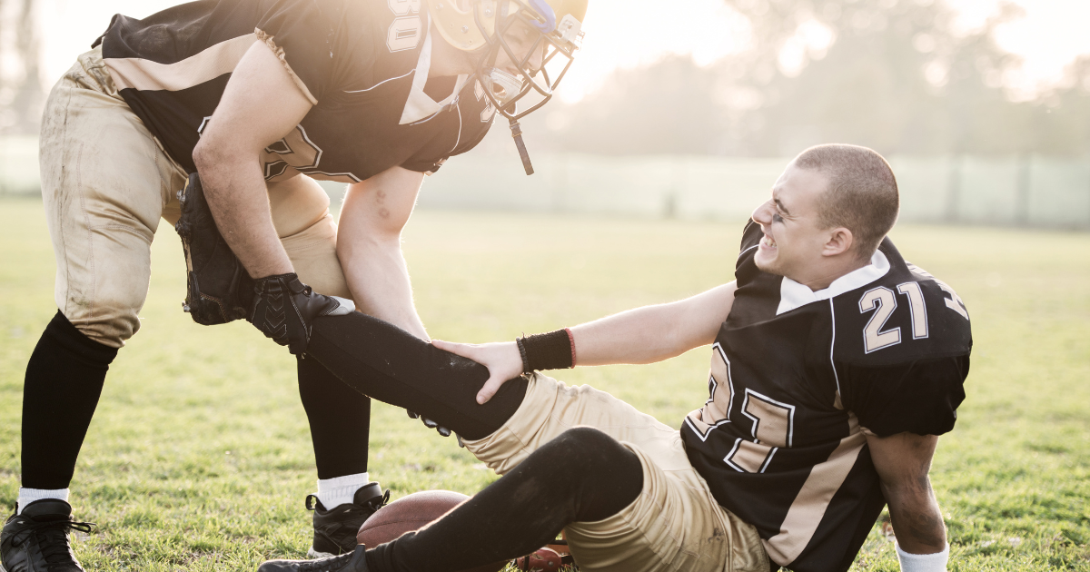 Football player on the ground, holding his leg, being assisted by a teammate. Both wear black and gold uniforms on a grassy field.
