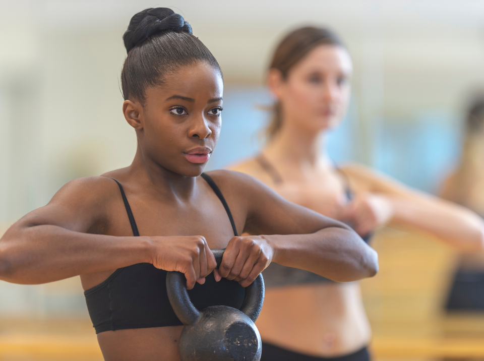 Woman lifting a kettlebell in a fitness studio, with another person exercising in the background.