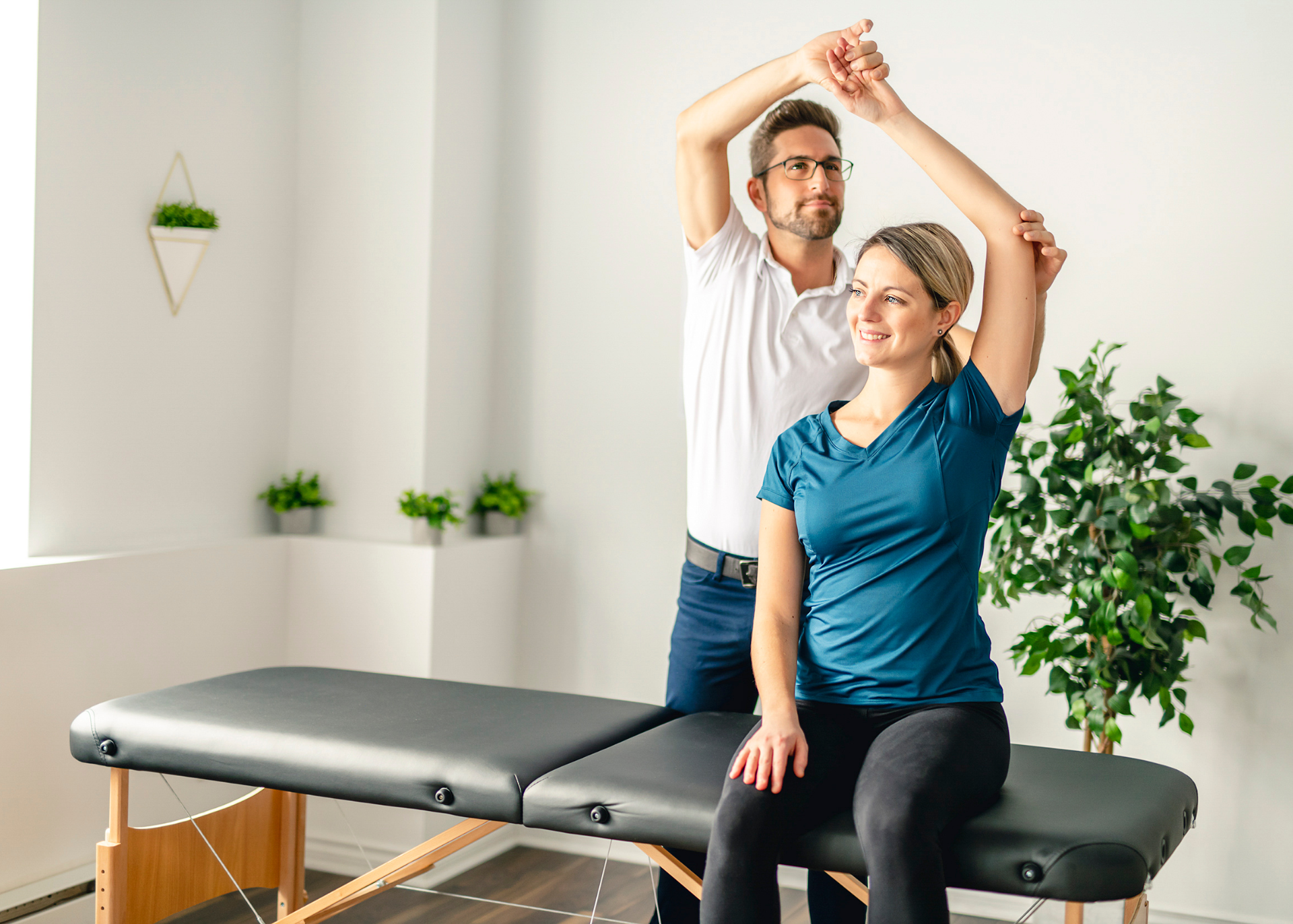 Physical therapist assisting a woman with arm movement in a well-lit room. The woman smiles while seated on a massage table.