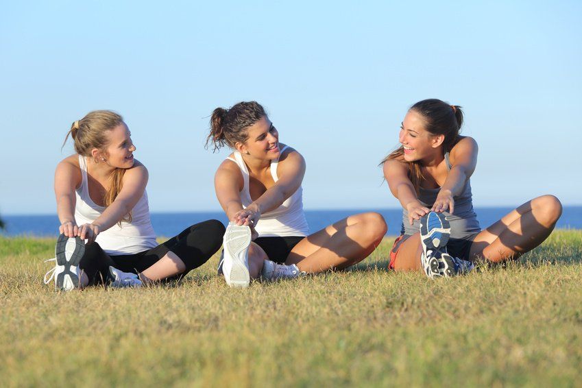 Three people sitting on grass outdoors, stretching and chatting under a clear blue sky.