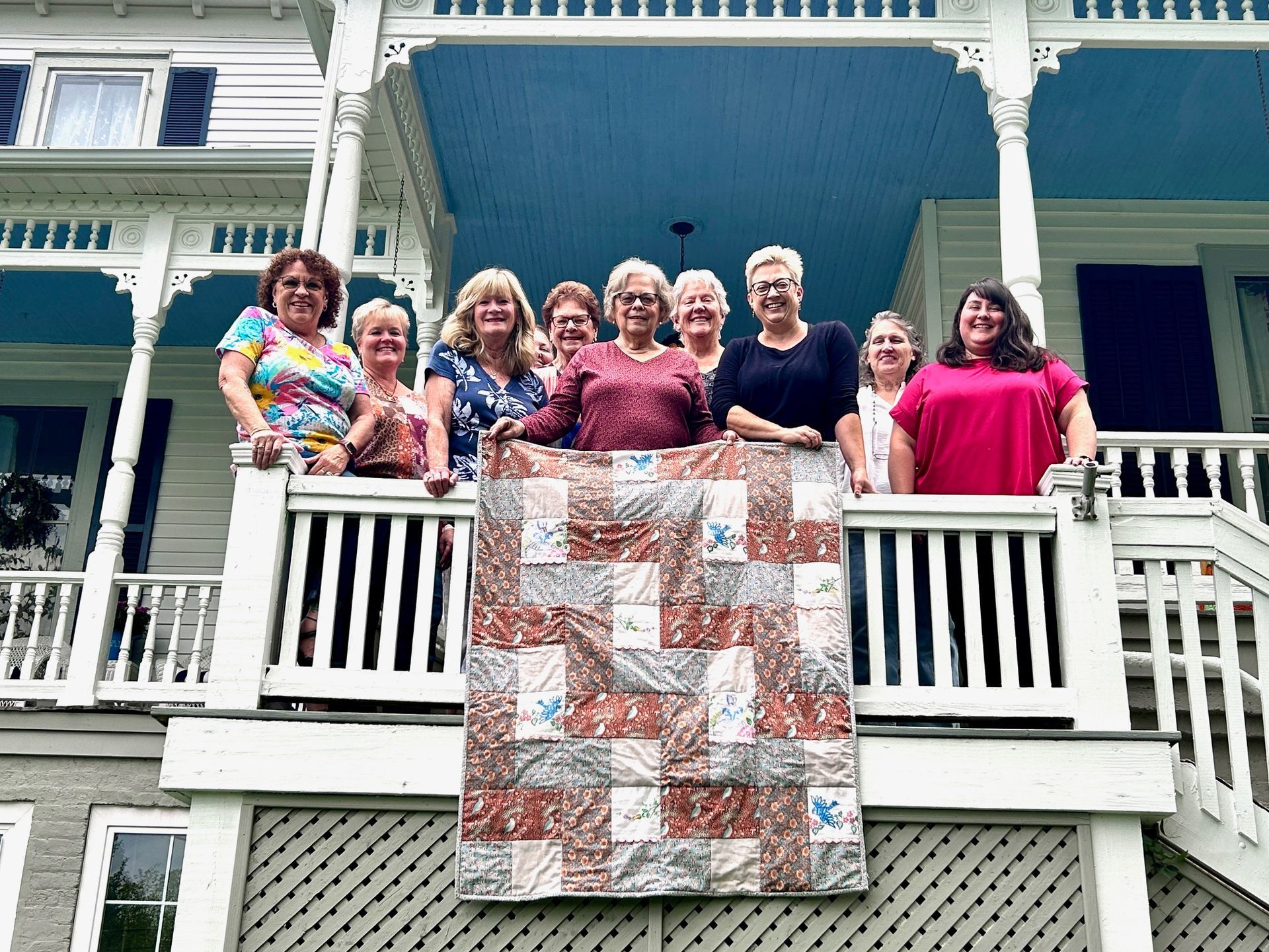 A group of women are standing on a porch holding a quilt.