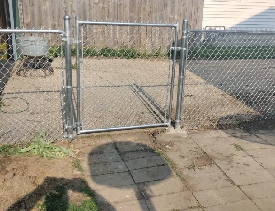 Chain-link fence gate in a backyard, with a shadow on paving stones and a wooden fence behind it.