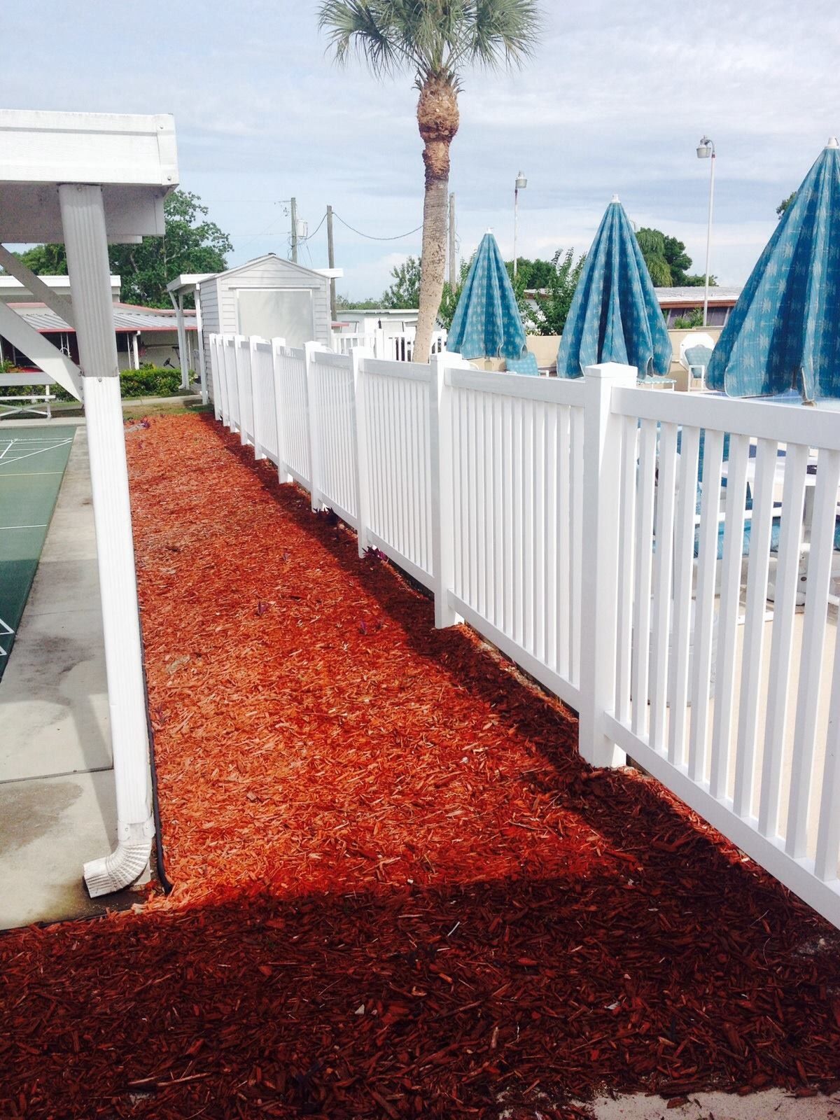 Colorful red gravel walkway beside a white fence, leading to a pool area with palm trees and blue umbrellas.