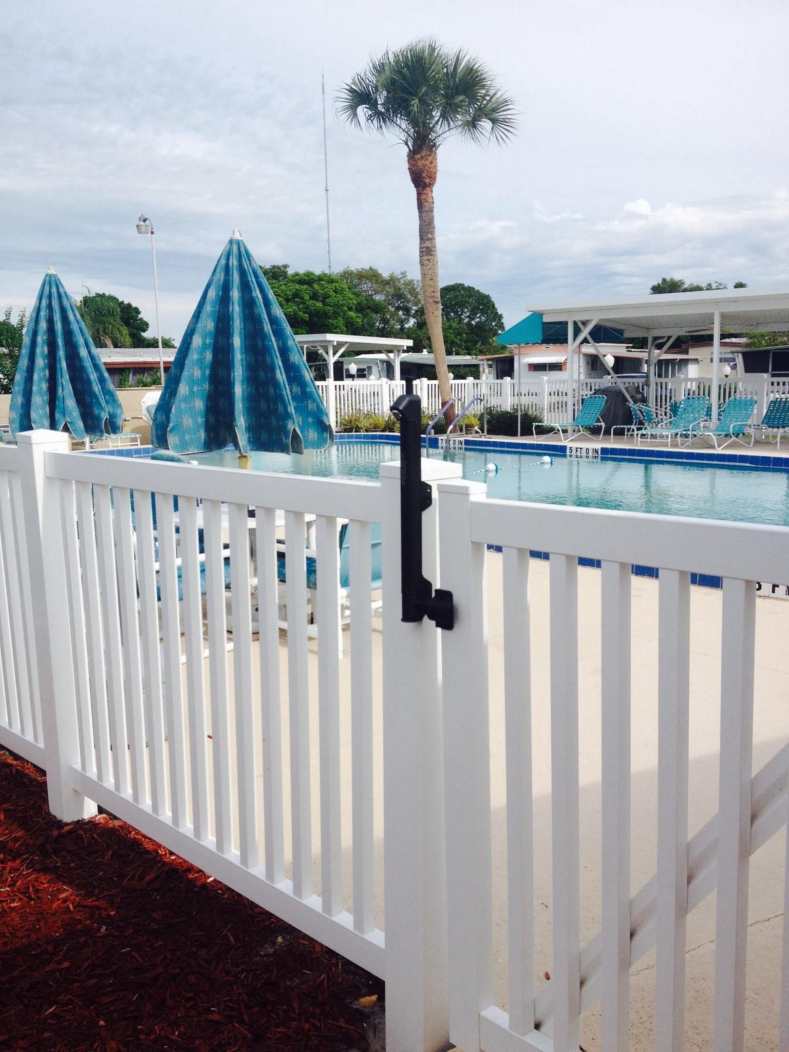 White fence by a pool with blue umbrellas and a palm tree under a cloudy sky