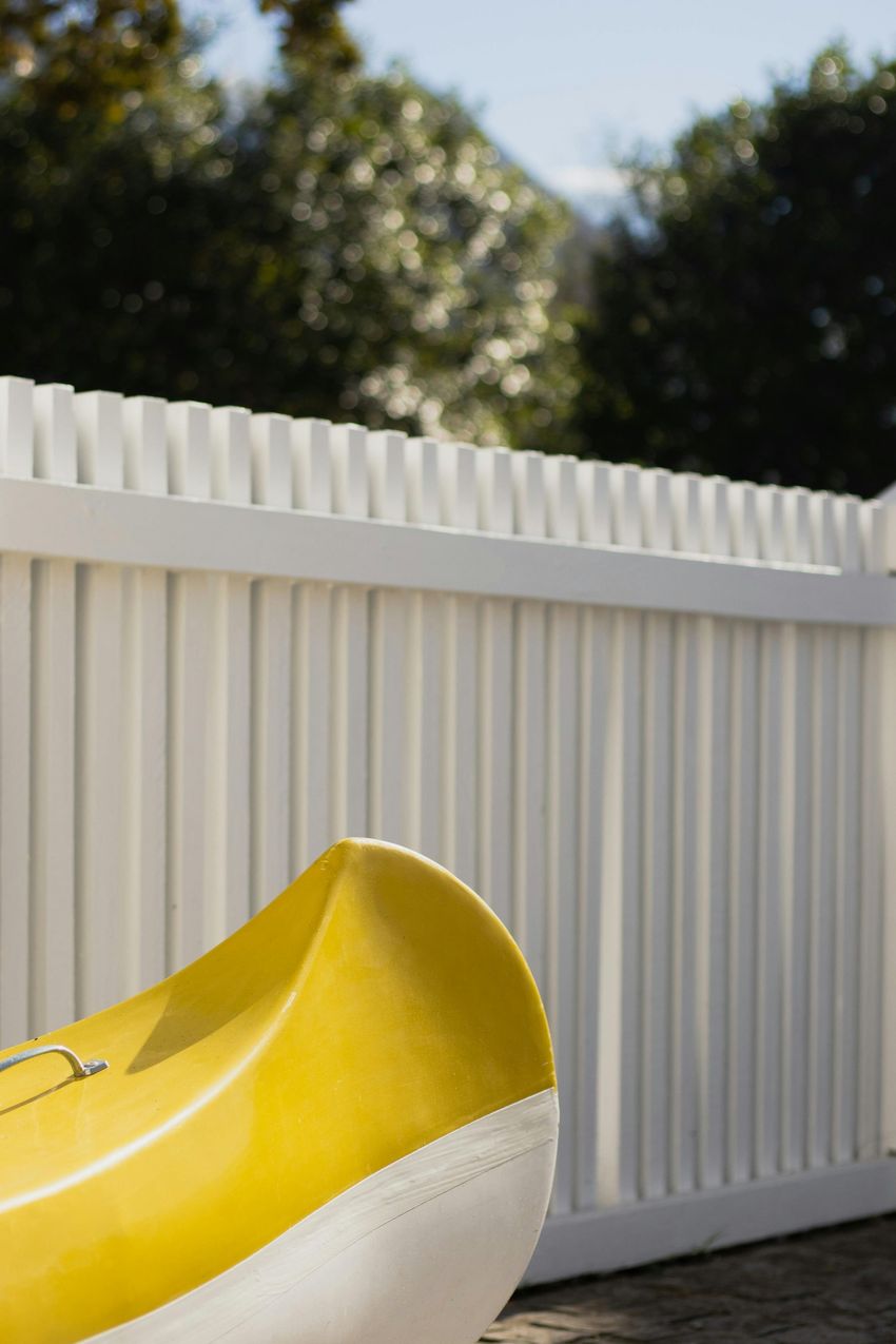 Yellow kayak beside a white vinyl fence with trees in the background