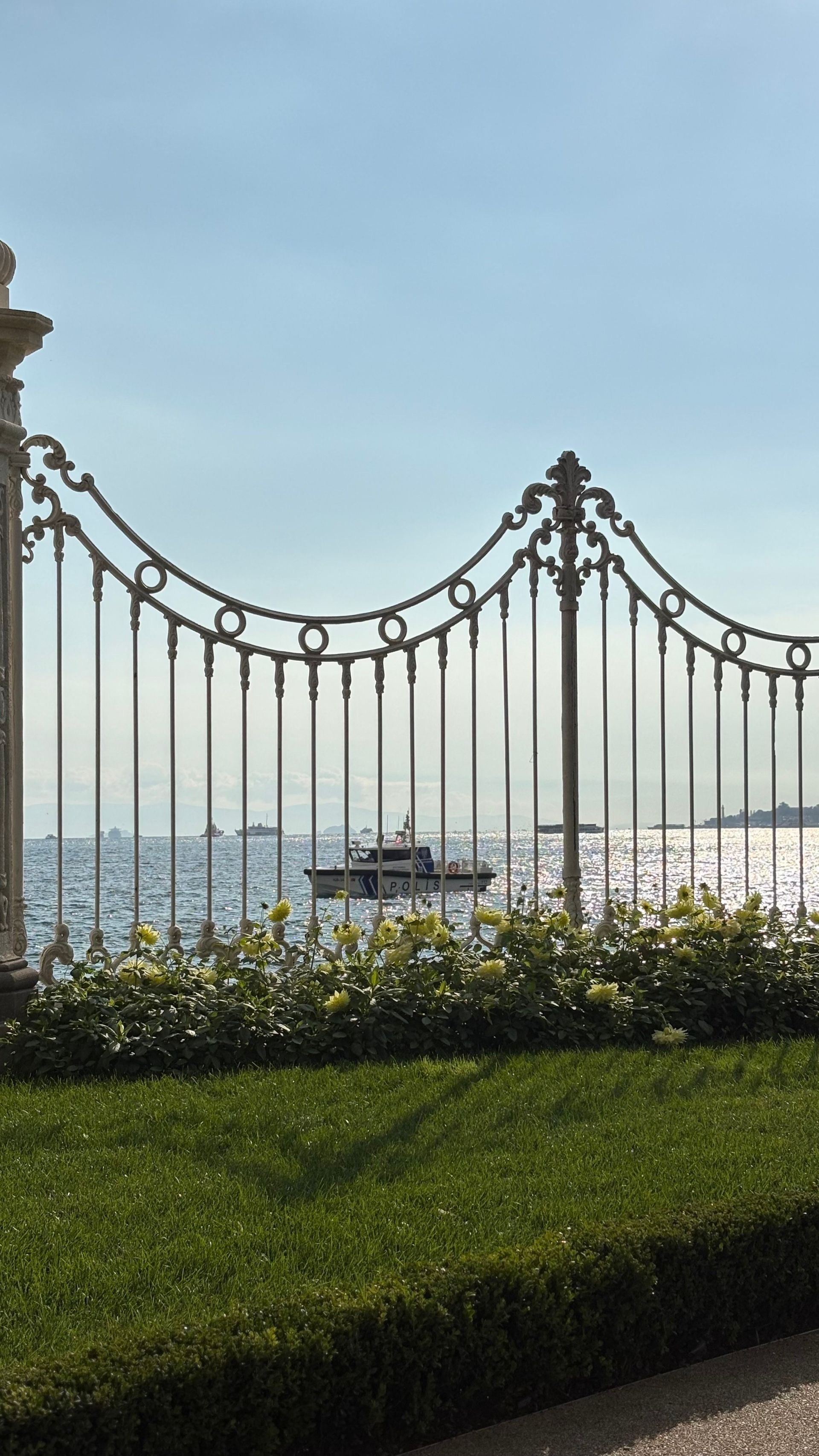 Harbor view through ornate iron fence, with boats on calm water and a green hedge in the foreground