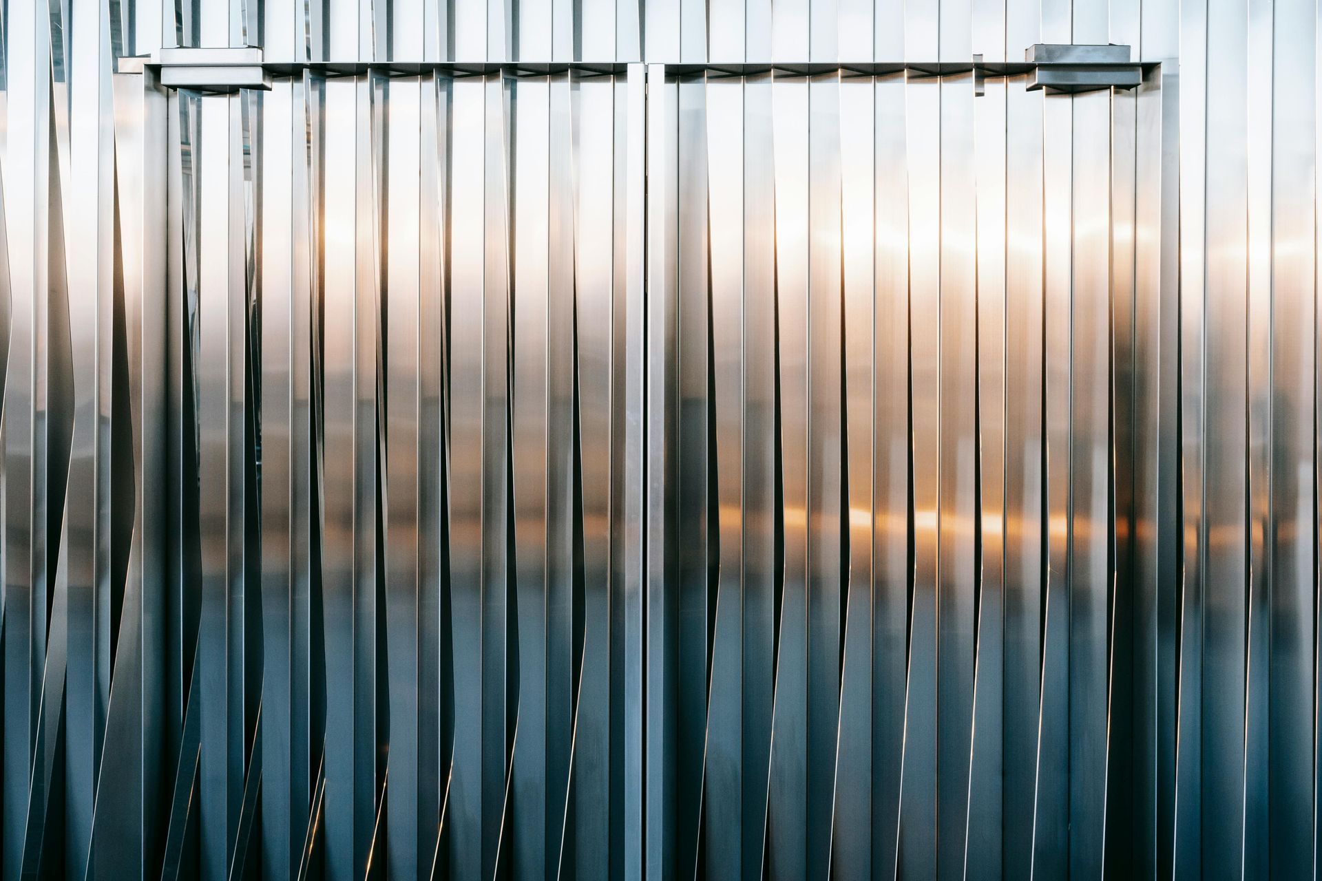 Vertical metallic fence or blinds with reflective silver-blue panels and warm light behind them