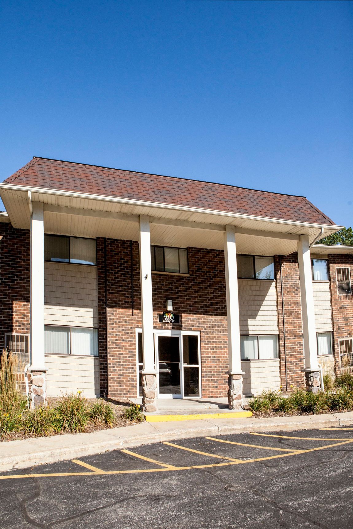 Apartment building entrance with brick facade, white columns, and a brown tiled roof under a clear blue sky.