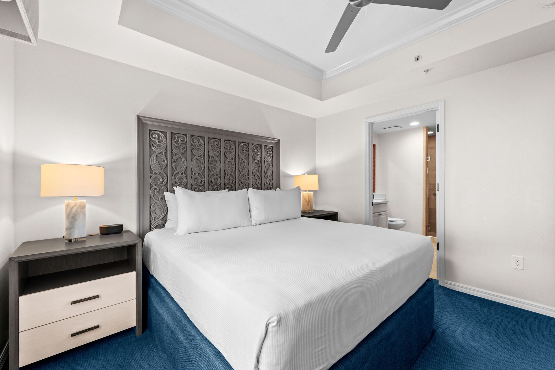 Bright hotel bedroom with king bed, blue carpet, nightstands, lamps, and patterned headboard.