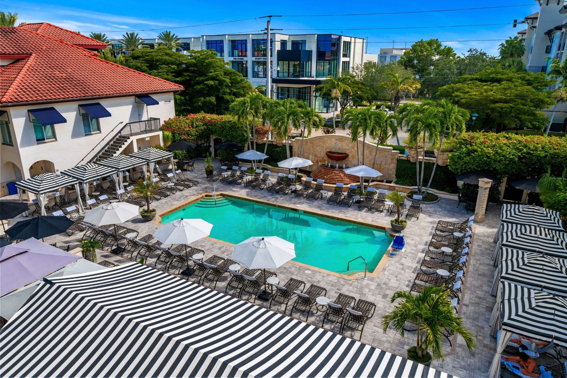 Resort pool with striped umbrellas, lounge chairs, and modern buildings under a bright blue sky