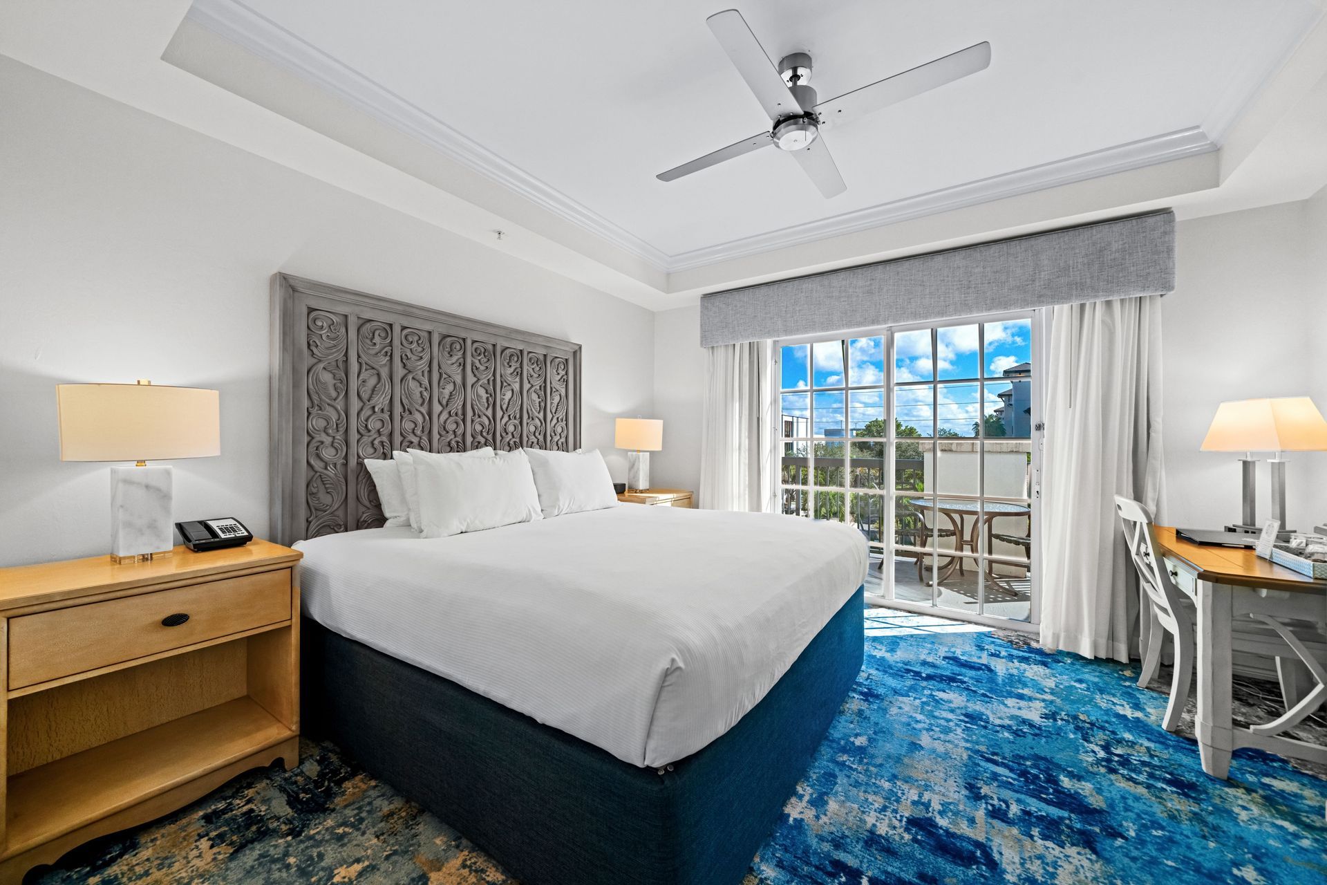 Bright bedroom with a white bed, blue rug, balcony doors, and a desk by the window.
