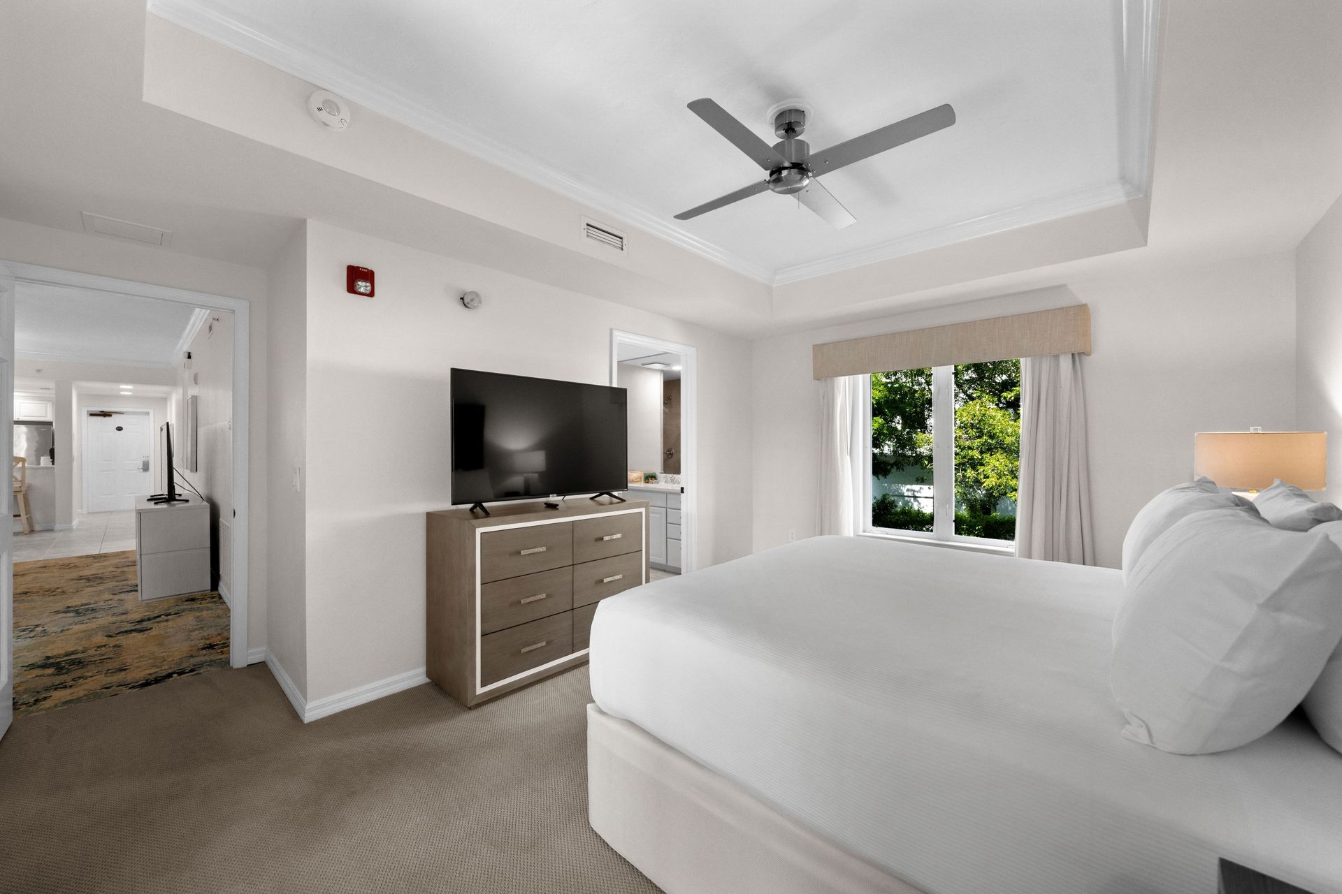 Bright bedroom with white bed, dresser, ceiling fan, and window overlooking greenery