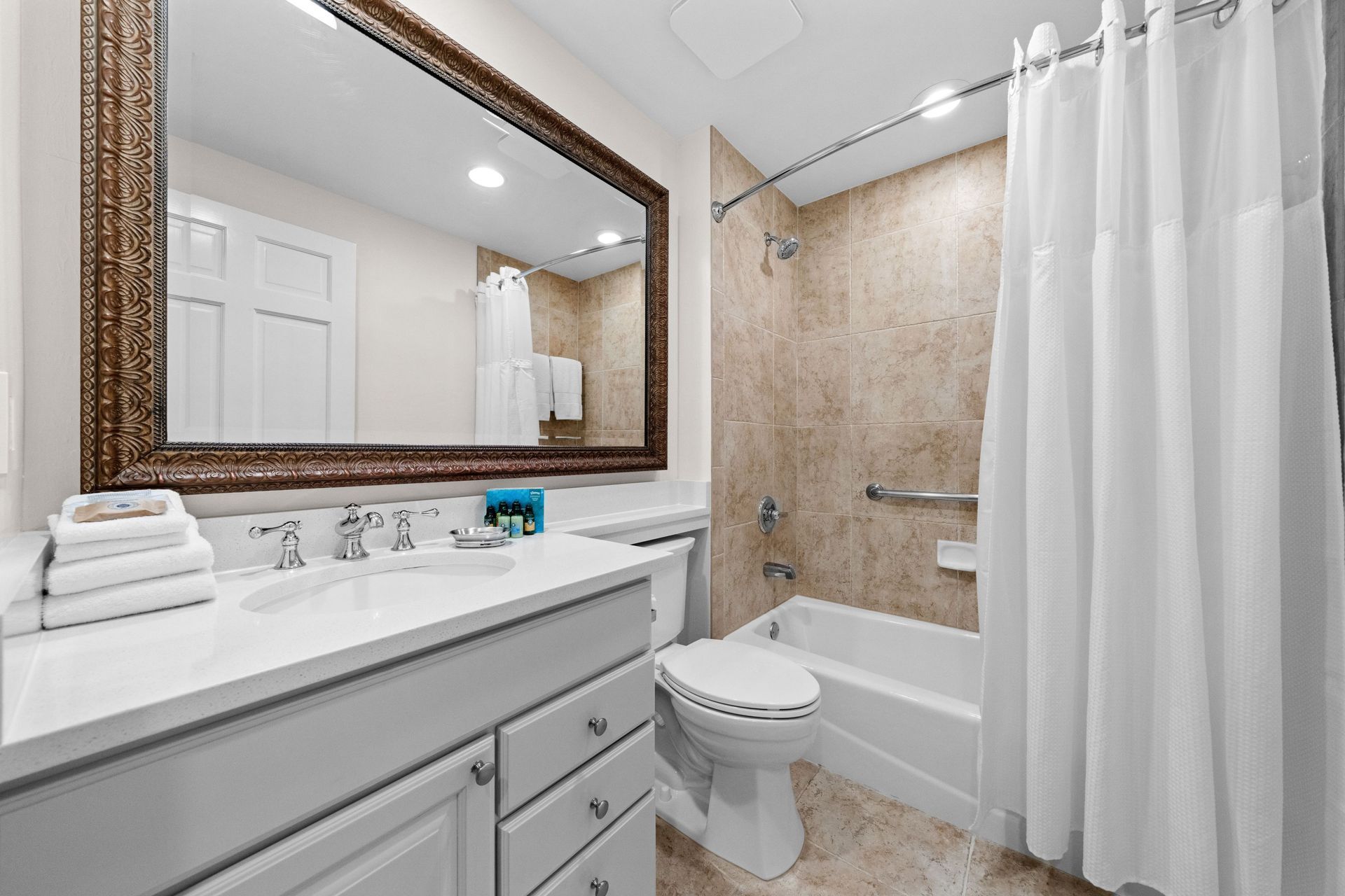 Bright bathroom with vanity mirror, white tub, beige tile shower, toilet, and white curtain.