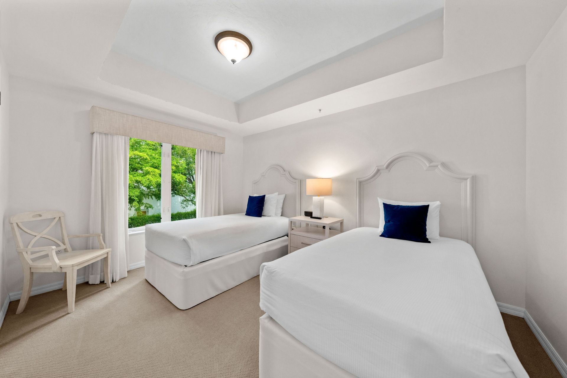 Bright twin bedroom with two white beds, blue pillows, and a window overlooking greenery