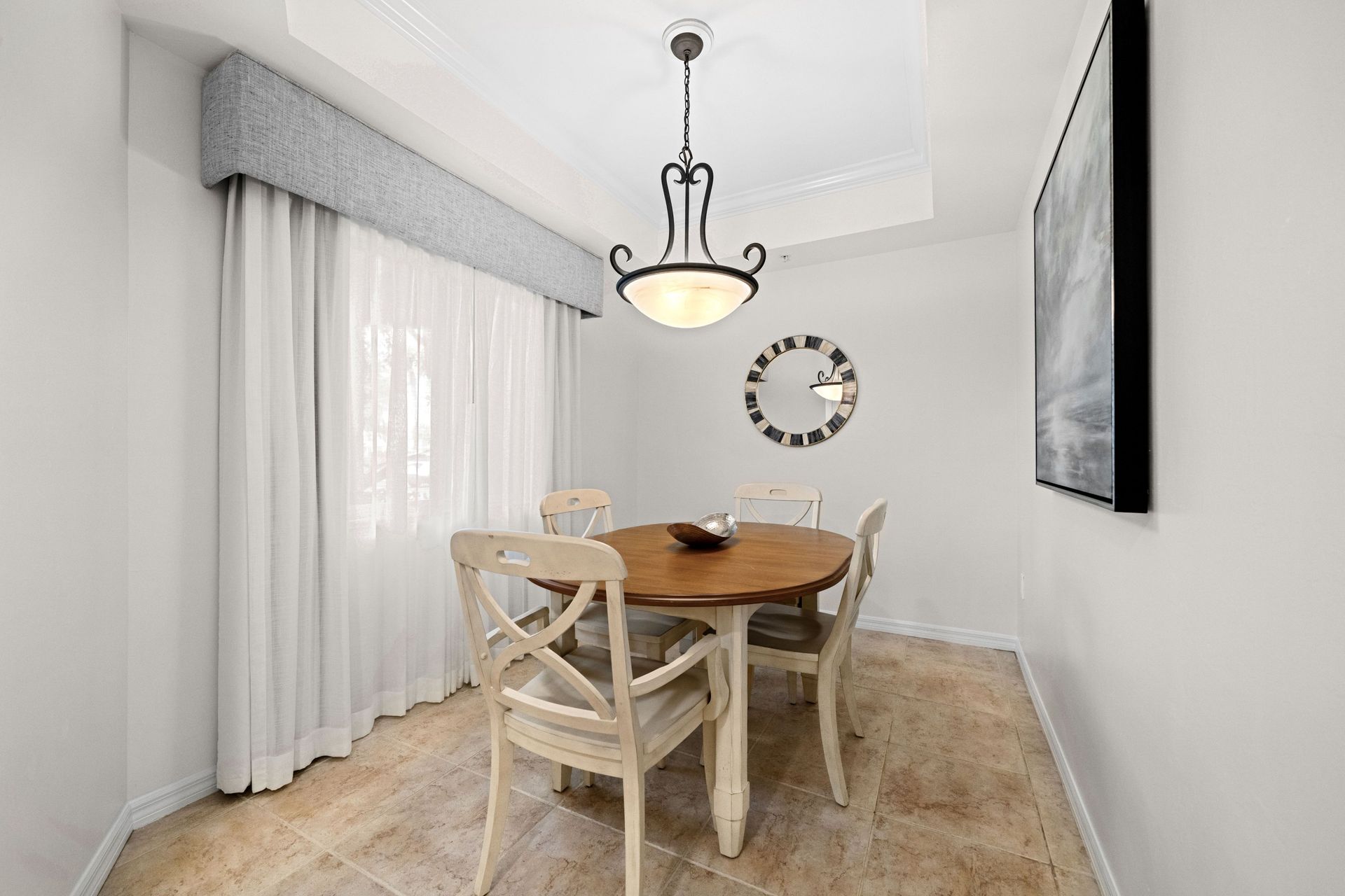 Bright dining room with a round wooden table, two chairs, chandelier, sheer curtains, and wall mirror.