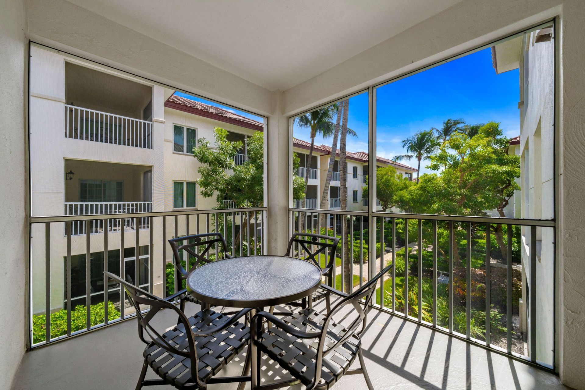 Balcony with round metal table and chairs overlooking apartment courtyard with green trees and blue sky