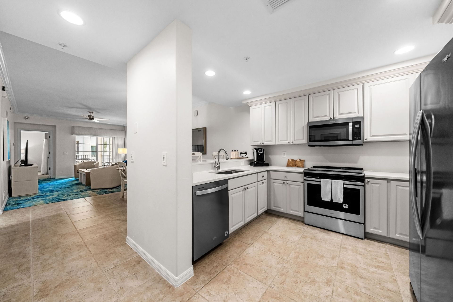 Modern kitchen with white cabinets, stainless steel appliances, and beige tile floors opening to a living area
