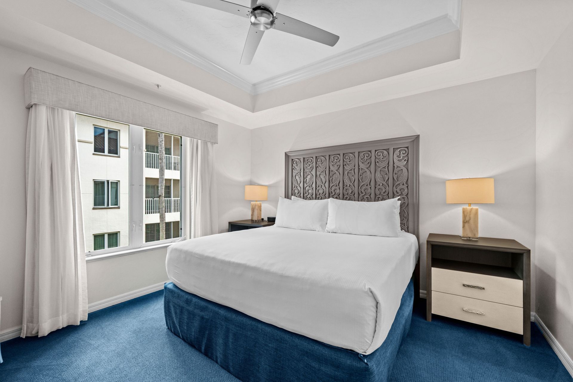 Modern bedroom with a blue bedspread, patterned headboard, and two bedside lamps by a window.