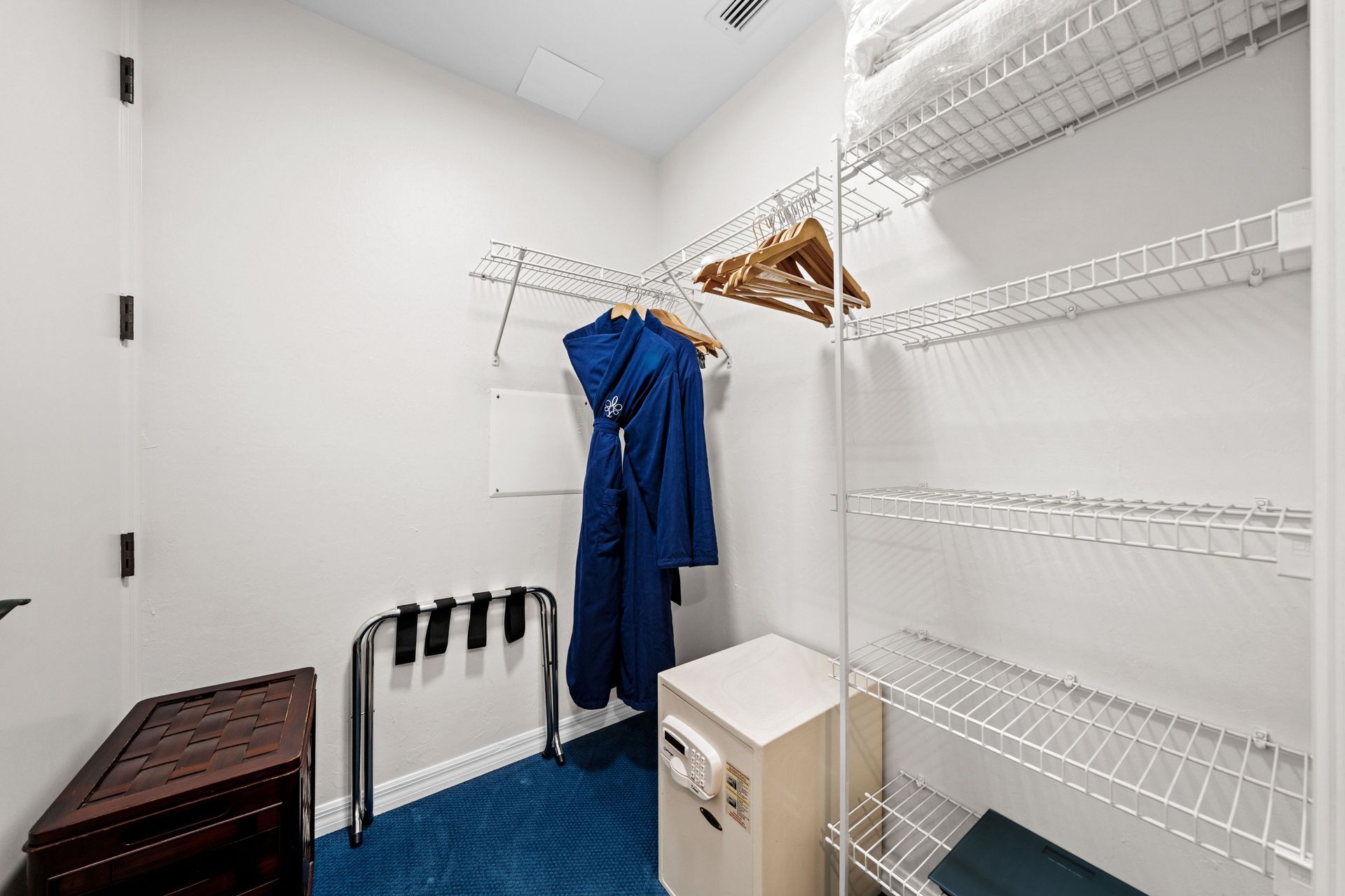 Small white closet with wire shelves, hanging blue coats, a white cabinet, and a blue carpeted floor.