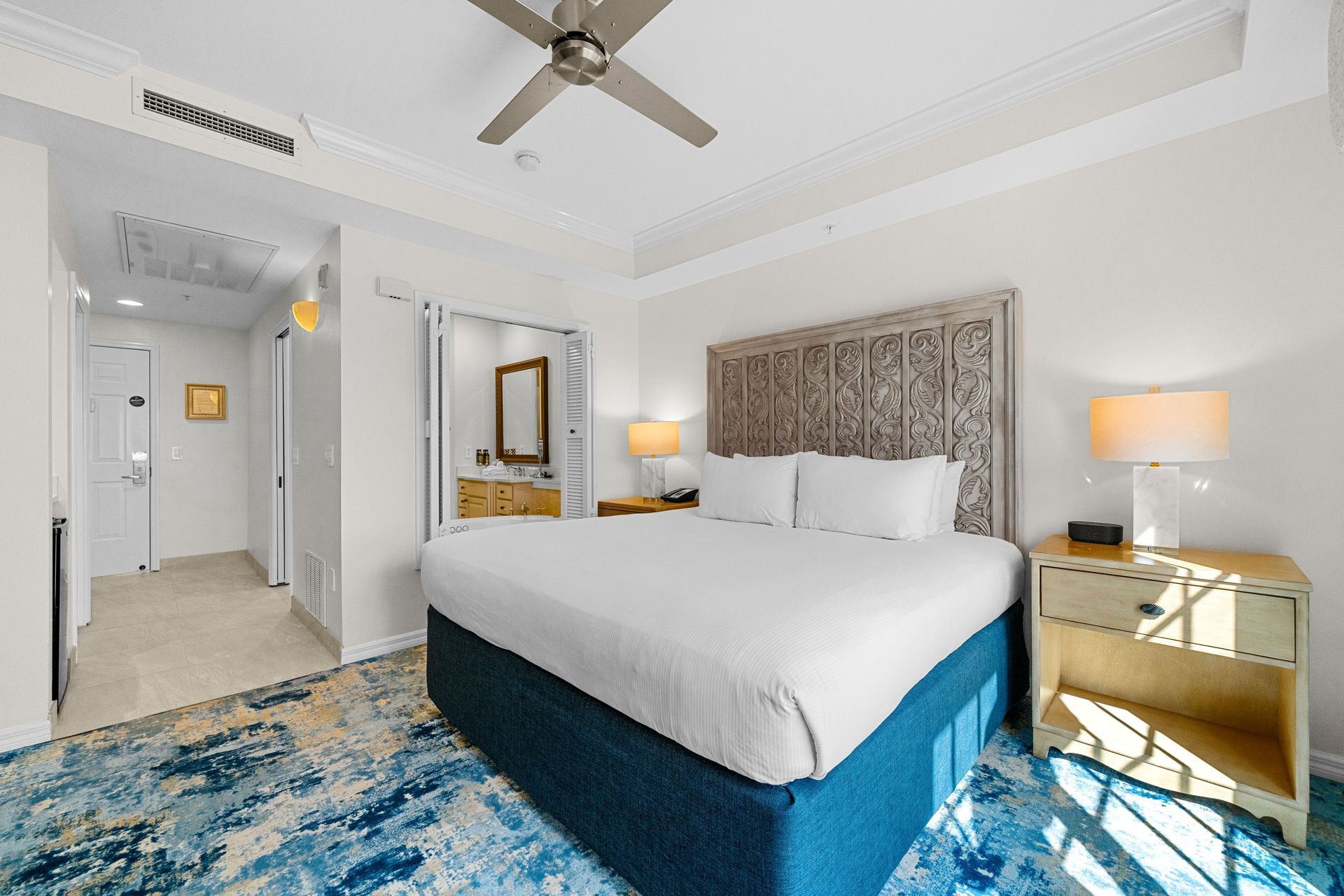Bright hotel bedroom with king bed, blue rug, nightstands, lamps, and ceiling fan