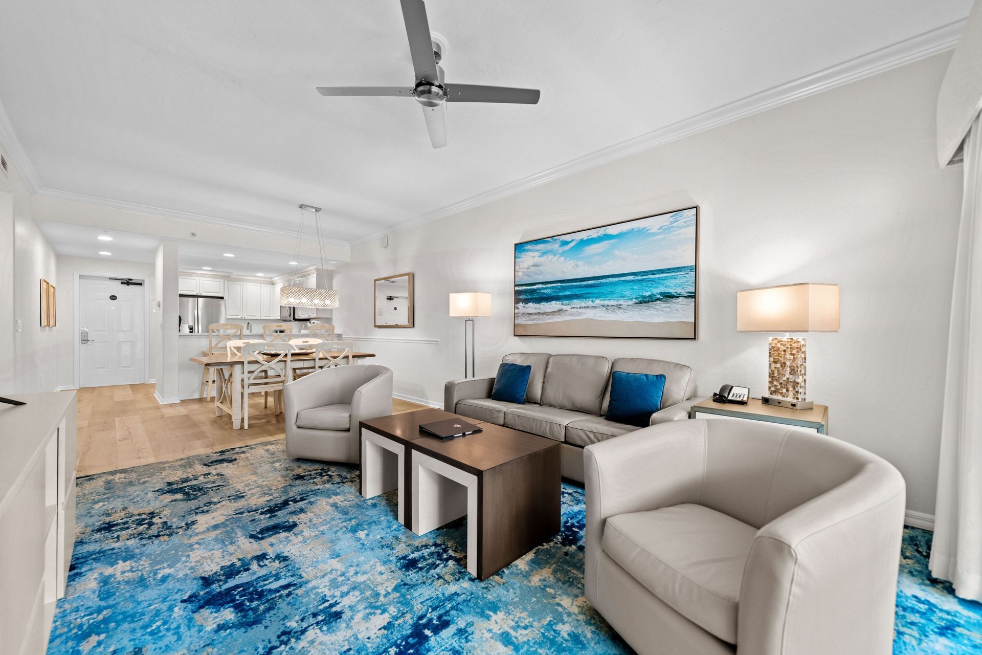 Bright modern living room with beige chairs, blue rug, wall art, and open kitchen in the background